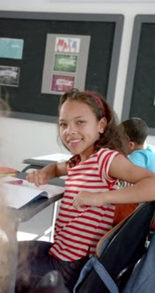 Vertical video: In school, girl smiling and sitting at desk in classroom, looking at camera