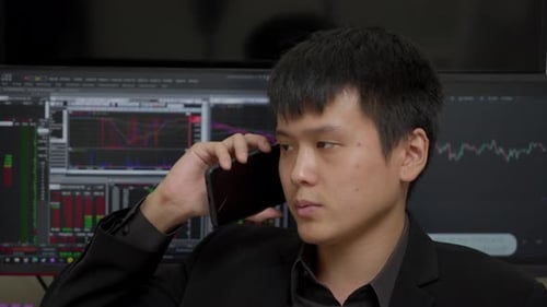 Chinese Businessman Talking On The Phone With Stock Market On Computer Monitors In Background. close