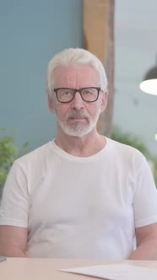Mature Man Wearing Glasses Sitting Indoors Looking at Camera