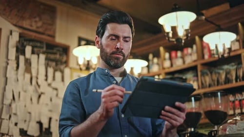 Focused Bearded Waiter Tapping with Stylus on Tablet While Standing at Bar