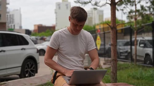 Indépendant masculin sérieux et prospère, tapant à distance, travaillant à distance sur un clavier d'ordinateur portable, assis sur un banc