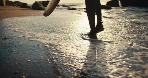 Nahaufnahme: Ein Mann in einem Neoprenanzug und Spezialschuhen hält ein Surfbrett in seinen Händen und geht den