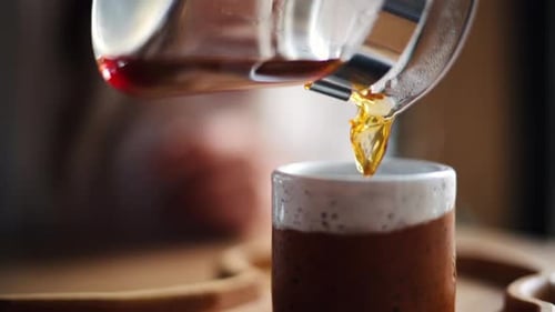 Close up of a woman pouring coffee in a cup from a glass pot standing on a wooden tray