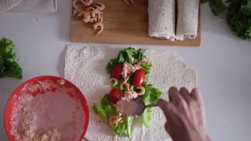 Weibliche Hände bereiten einen Wrap mit frischem Salat, Tomatensauce und Garnelen auf einem flachen Tortilla-Teil zu