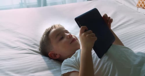 Cute Smiling Child Kid Boy Using Digital Tablet Technology Device Lying on Bedroom Alone