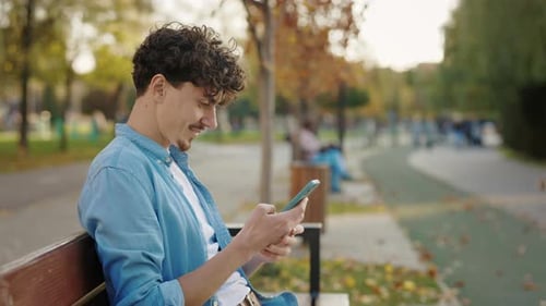 Side View of the Bearded Man Sitting on the Bench in the Street Using His Smartphone Texting With