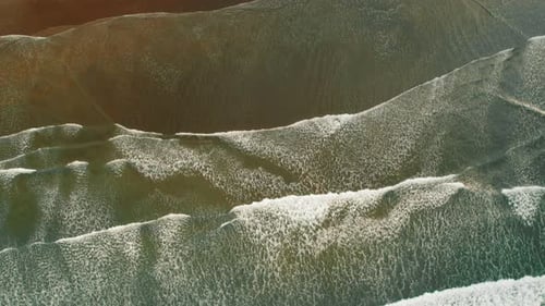 Top view of ocean waves rolling to dark sandy shore with textured white foam