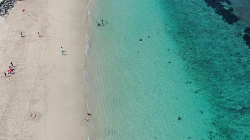 Drone captures an aerial image of the sea's azure water near a beach where people are strolling on t