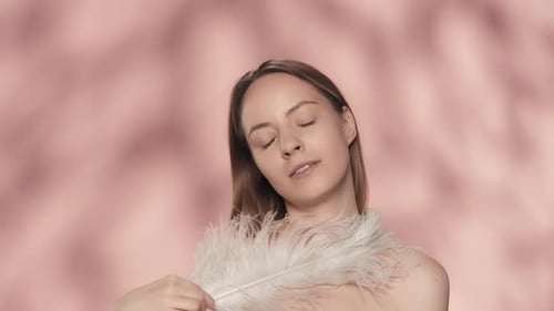 A Woman Touches Her Bare Shoulders and Face with a White Feather A Young Woman Demonstrates Smooth