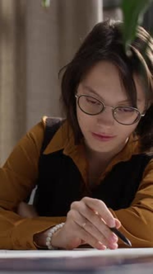 Vertical of Young Female Architect Analyzing Blueprint at Office Desk