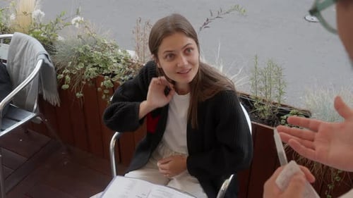 A young woman chats with waiter at summer cafe terrace and orders from the menu.