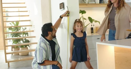 Pai de família ajoelhado estendendo fita amarela, medindo a altura da menina contra a parede na cozinha