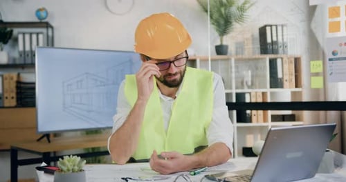 Man architect in vest and helmet posing on camera at his workplace with blueprint in design office