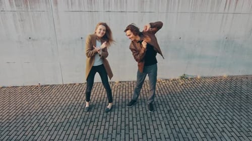 Cheerful Girl and Happy Young Man with Long Hair are Actively Dancing on a Street next to an Urban