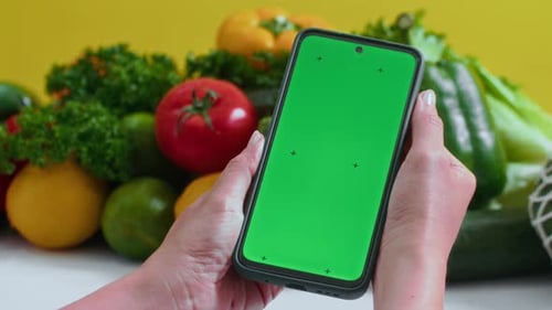 Hands of Person Holding Chroma Key Smartphone in front of Fresh Vegetables
