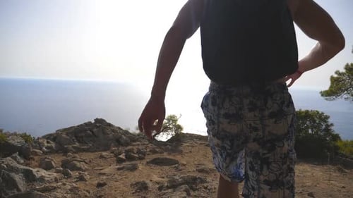Unrecognizable Hiker with Backpack Reaching Up Top of Mountain with Ocean at Background