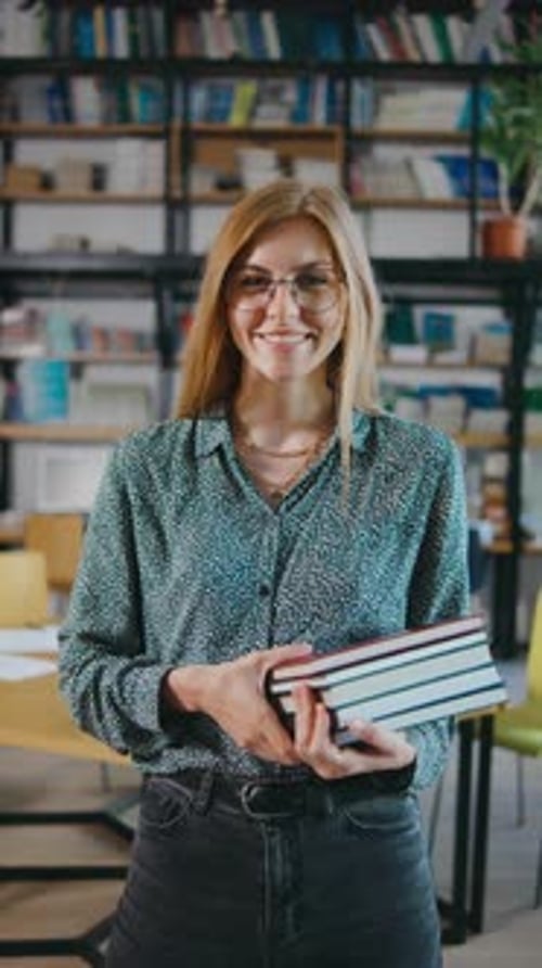 A Young Woman with Glasses in a Library Smiling Holding Books and Browsing Shelves Woman Holding a