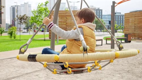 Slow motion of 7 year old boy swinging and spinning on the public playground on the windy sunny day.