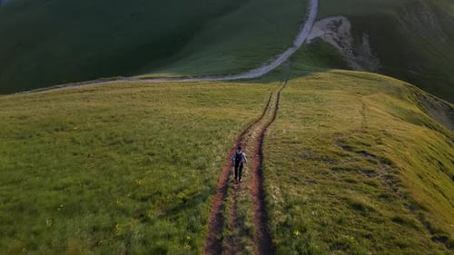 A man with a backpack walks along a green mountain path at sunset. Aerial view.