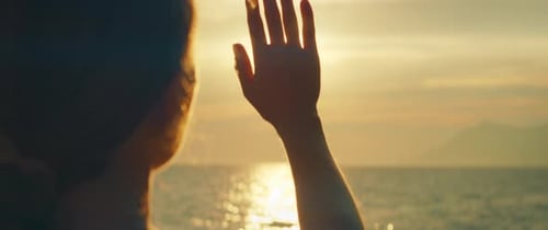 Person raising hand toward bright sunset over calm ocean horizon in the evening