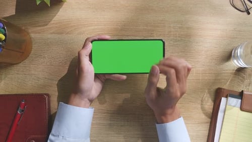 Top down view of man's hands using green screen smartphone on a wooden desk