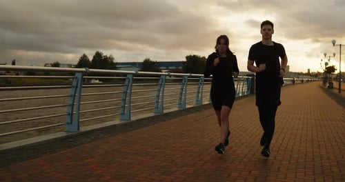 Young Man and Woman Running Along the Embankment in the City at Sunset