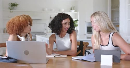 Trois femmes adultes assises à la table de la cuisine, femme centrale pointant vers un ordinateur portable, écrivant des notes sur une tâche