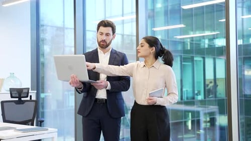 Two co-workers, man and woman engaged in discussion holding a laptop in office.