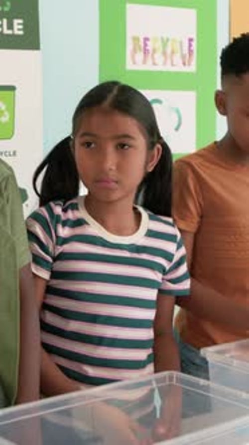 Vertical video: Adult offering recyclables, girl in green tee sorting paper into PAPER bin in class