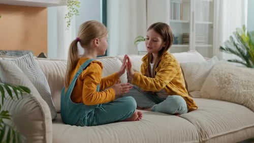 Two Young Girls Clapping Hands on Sofa Indoors