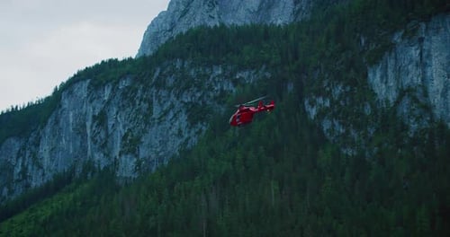 Red rescue helicopter flying over lush green Austrian mountain landscape with rocky cliffs
