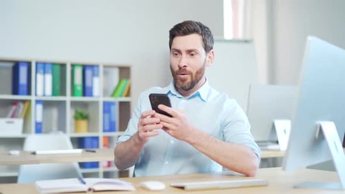 happy handsome office worker sitting at a computer desk in the office using a mobile phone. Cheerful