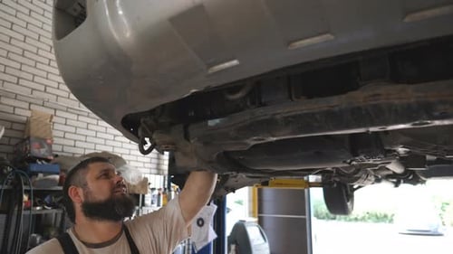 Professional Bearded Auto Mechanic Working Underneath a Lifting Vehicle at Service or Garage
