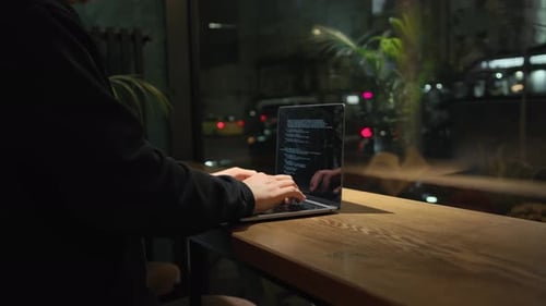 A man is programming, typing a code on a laptop at night in an institution, possibly a hacker