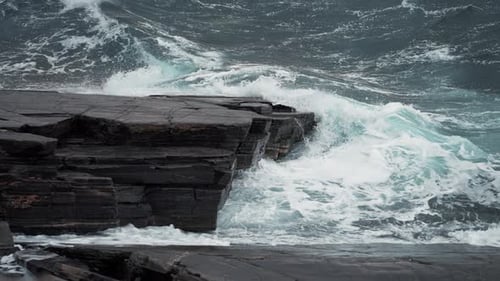 Heavy waves hit the dark jagged rocks on the shoreline. Turquoise water foaming and raging. Slow-mor
