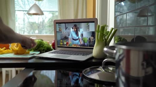 Una mujer sigue un tutorial de cocina en línea en una computadora portátil mientras prepara alimentos saludables en casa