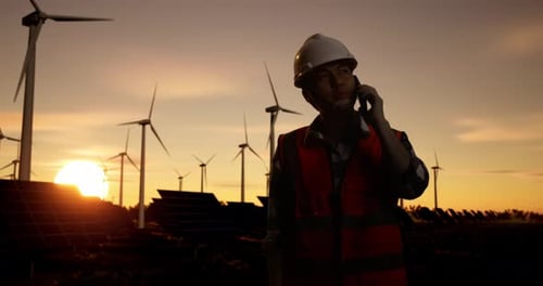 Male Engineer In A Helmet Talking On Smartphone And Looking Around With Wind Turbines At Sunset