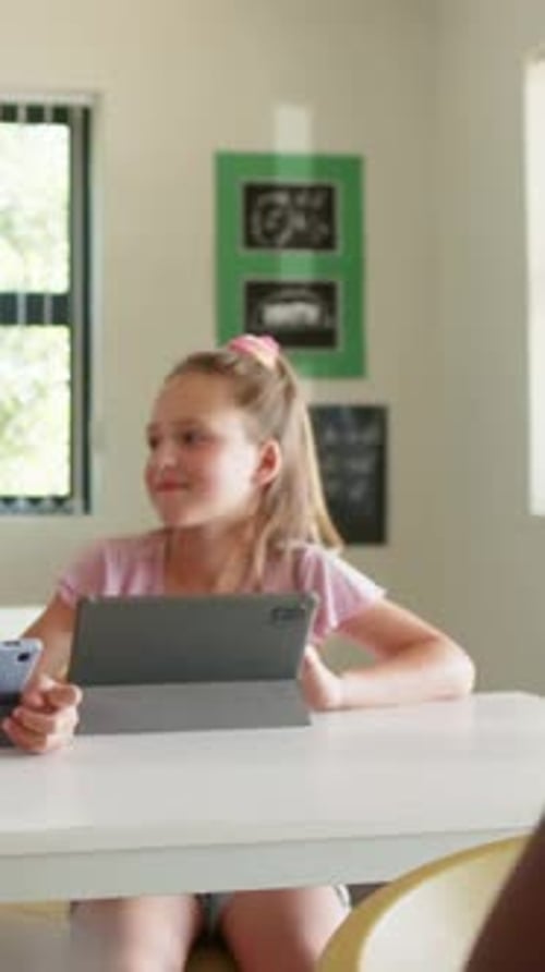 Vertical video: Reacting to peer entering left, pink top girl turning head in class with tablet