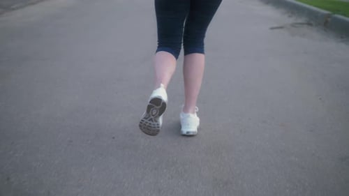 Nahaufnahme der Beine einer Frau, die auf einem betonierten Fußweg im öffentlichen Park mit Sonnenlicht im Hintergrund läuft