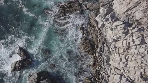 Descending rotating birdseye aerial view above rocky beach splashing rough blue sea waves shoreline