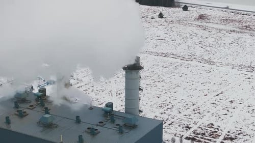 Industrial smokestacks emitting steam against a snowy landscape, overcast weather, aerial view