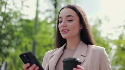 Smiling Female Using Modern Gadget While Standing in Park and Holding Coffee Cup