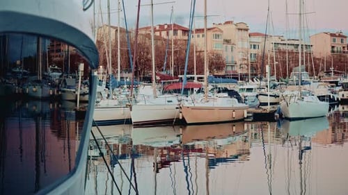Sailboats and yachts docked in a calm marina, reflected in still water with pastel colored residenti