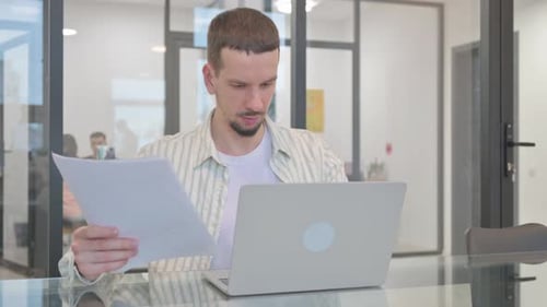 Man Works on Laptop and Reviews Documents in Office