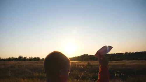 Little Boy Holding in Hand Paper Airplane and Walking Through Grass Field