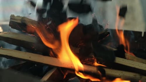Chunks of Black Charcoal Falling in Slow Motion Onto Burning Firewood in a Grill Creating Sparks and