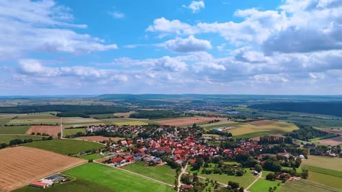 A rural village surrounded by wide multi-colored fields under a cloudy sky.