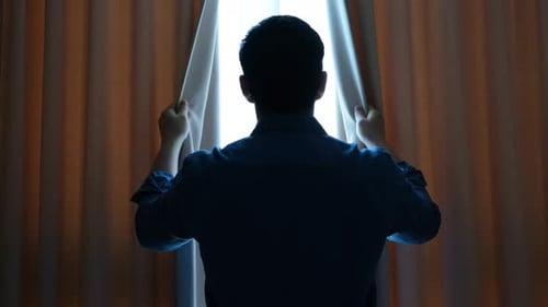 Young man in blue shirt looks out the window and opens the curtains, rear view.