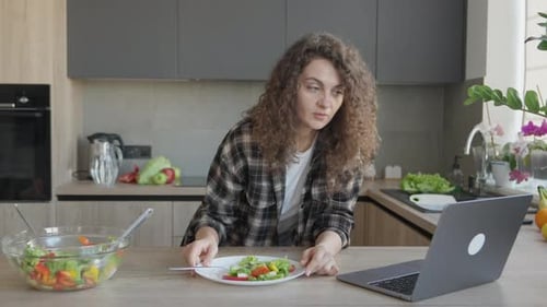 Woman Eating Salad And Smiling During Video Call In Bright Modern Kitchen