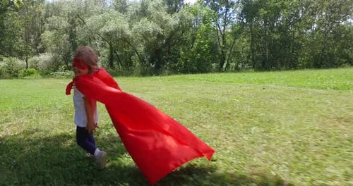 a little girl dressed as a comic book hero runs happily in a beautiful garden . sunny day and hap